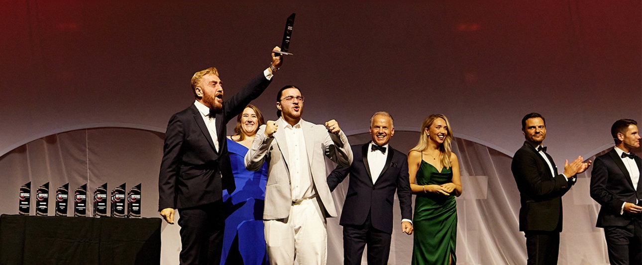Herr Göksel steht als Gewinner des Mazda-Awards auf der Bühne und zeigt 
stolz seinen Pokal - neben ihm elegant gekleidete Menschen.
